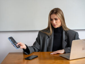 Na pierwszym planie obrazka znajduje się biurko na, którym leży telefon i laptop. Na drugim planie znajduje się kobieta siedząca przy biurku trzymając telefon w dłoni.
