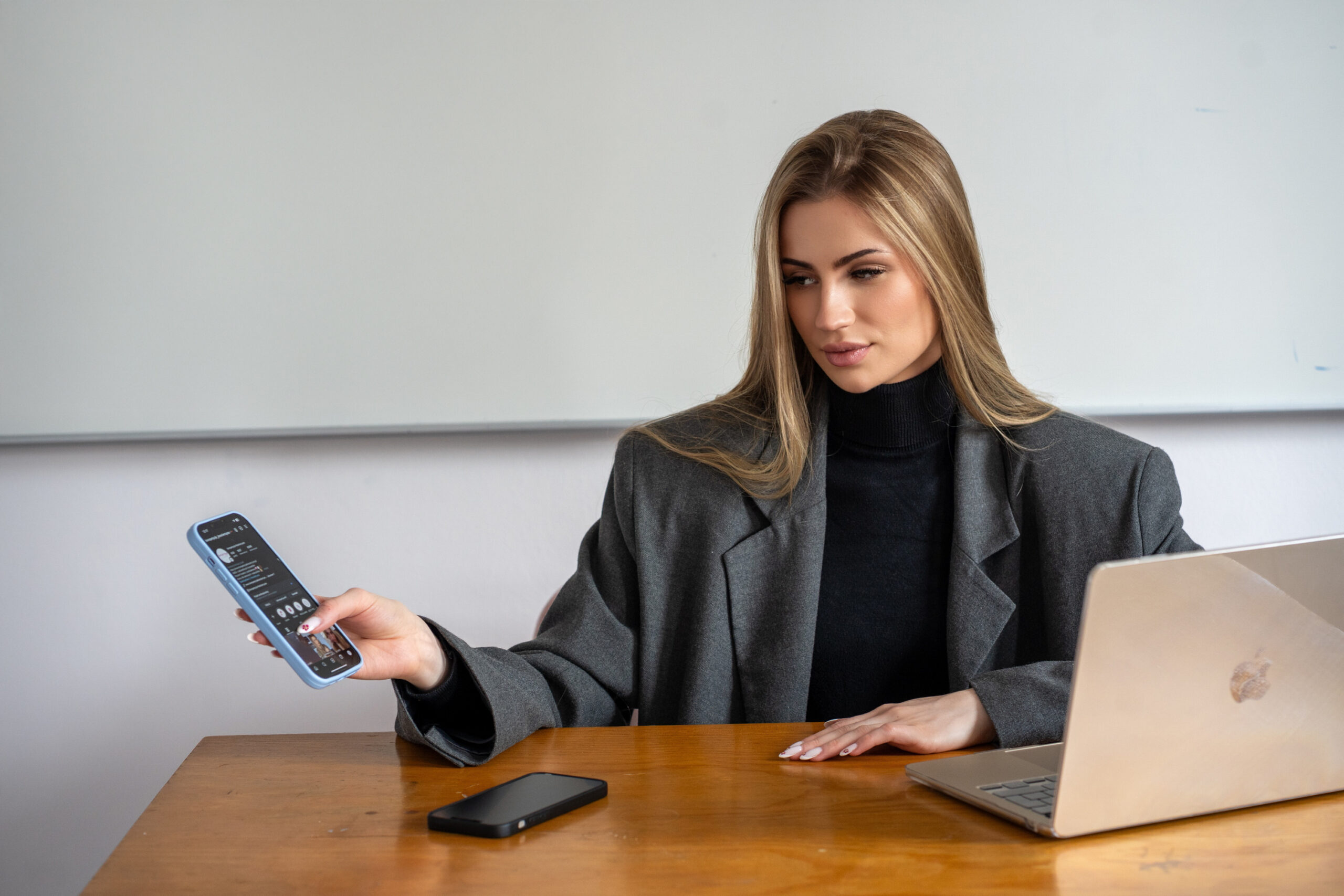 Na pierwszym planie obrazka znajduje się biurko na, którym leży telefon i laptop. Na drugim planie znajduje się kobieta siedząca przy biurku trzymając telefon w dłoni.